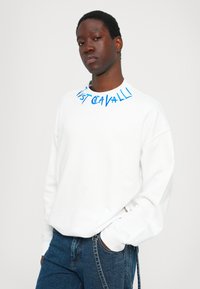 Λευκή μπλούζα με μπλε γράμματα "Just Cavalli" γύρω από τον λαιμό, χαλαρός στυλ, μακριά μανίκια και  ribbed μανσέτες. Ορατό τζιν παντελόνι από κάτω.
