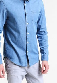 Camicia in denim azzurro chiaro con maniche lunghe, colletto con bottoni e tasca sul petto. Presenta bottoni bianchi e un orlo curvo.