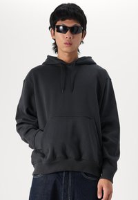 Sweat-shirt à capuche noir avec une poche kangourou, des poignets et un ourlet côtelés, fabriqué en tissu doux. Porté avec des lunettes de soleil noires.