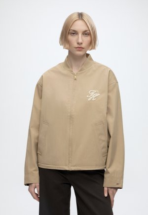 Donna con capelli biondi corti che indossa una giacca beige con zip e un logo ricamato sul petto e pantaloni neri, rivolta in avanti.