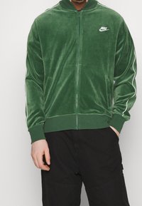 Green velvet zip-up jacket with white side stripes, ribbed cuffs, and collar. Features a small white Nike logo on the left chest.