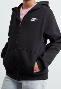 Persona con una sudadera negra con cremallera de Nike sobre una camiseta rosa, con las manos en los bolsillos delanteros y jeans de luz azul.