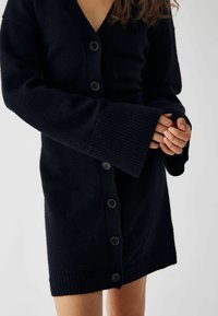 Melns, adīts kardigans ar V veida kakla izgriezumu, ribotām aprocēm un pogām pa priekšu. Iespējama teksturēta auduma un vidukļa garums.