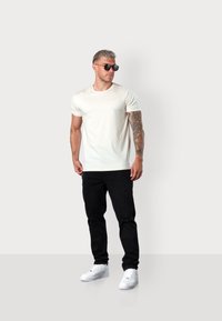 Hellbeige Kurzarm-T-Shirt kombiniert mit schwarzen, ausgebleichten Jeans und weißen Sneakers. Eine minimalistische Sonnenbrille wird getragen.