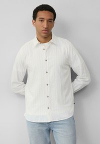 Witte blouse met verticale lichtblauwe strepen, kraag en bruine knoppen. Draagt bij lichte spijkerbroek met een gebogen zoom.