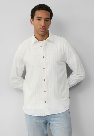 Chemise blanche à boutons avec des rayures verticales bleu clair, col et boutons marron. Portée avec un jean bleu clair, doté d'un ourlet arrondi.