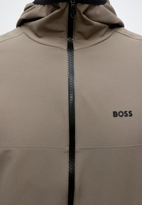 Μπεζ μπουφάν με μαύρο λογότυπο BOSS στο στήθος, κεντρικό μαύρο φερμουάρ με υφή και κουκούλα.