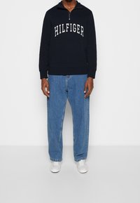 Felpa blu navy con zip e logo "HILFIGER", abbinata a jeans larghi azzurri e sneaker bianche, esposta su uno sfondo neutro.