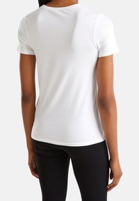 En person klädd i en enkel vit t-shirt med kort ärm och svarta byxor, sedd bakifrån mot en vit bakgrund.