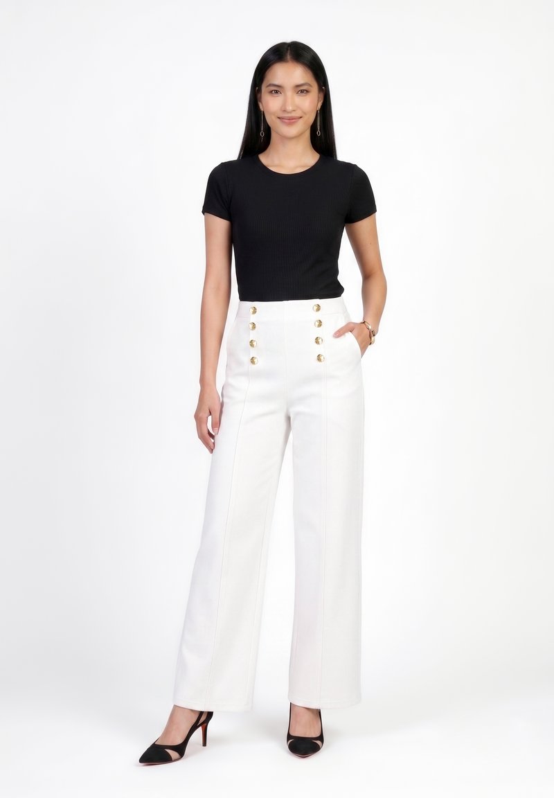 Femme debout portant un top noir à manches courtes, un pantalon blanc taille haute avec des boutons dorés et des talons noirs pointus sur fond blanc.