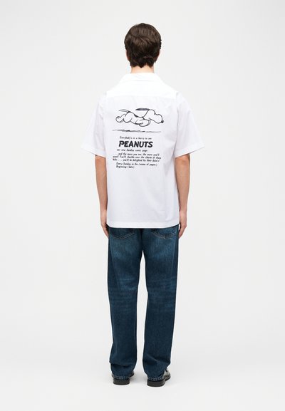 Άνδρας που φοράει λευκό κοντομάνικο πουκάμισο με εικονογράφηση του Snoopy και το κείμενο Peanuts στην πλάτη, συνδυασμένο με μπλε τζιν και μαύρα παπούτσια.