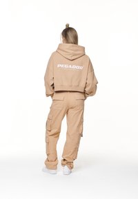 Sudadera corta beige con texto "PEGADOR", combinada con pantalones cargo que cuentan con bolsillos laterales y un corte holgado, complementados con zapatillas blancas.