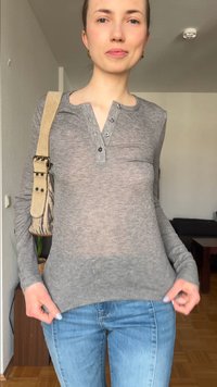 Nő szürke henley ingben és kék farmerben, beltéren áll, bal vállán bézs színű válltáskával.