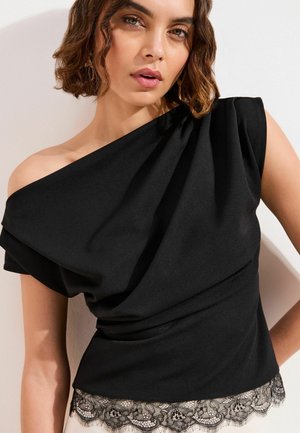 Kobieta z krótkimi, falowanymi włosami, ubrana w czarny top na jedno ramię z drapowanym materiałem i koronkową lamówką na dole, na jednolitym tle.