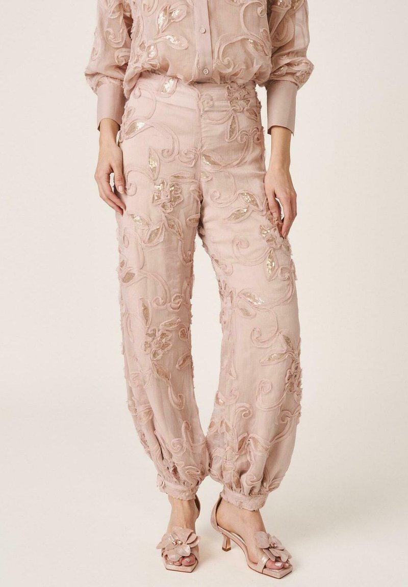 Pantalones de talle alto en rosa claro con bordados florales y adornos, combinados con blusa transparente a juego y sandalias de tacón rosas con detalle floral.