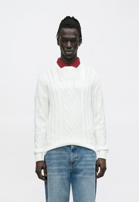 Młody mężczyzna o ciemnej karnacji i dredach, ubrany w biały sweter w splot kablowy z napisem "GUESS" na czerwonej koszuli z kołnierzykiem i niebieskich dżinsach.