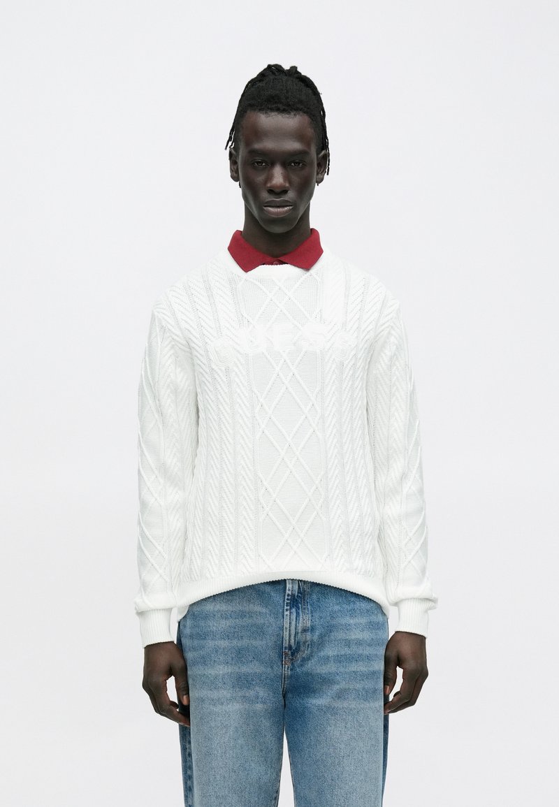 Młody mężczyzna o ciemnej karnacji i dredach, ubrany w biały sweter w splot kablowy z napisem "GUESS" na czerwonej koszuli z kołnierzykiem i niebieskich dżinsach.