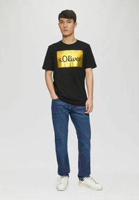 Schwarzes T-Shirt mit einem großen goldenen rechteckigen Patch, auf dem "s.Oliver." steht. Kombiniert mit blauen Jeans und weißen Sneakern.