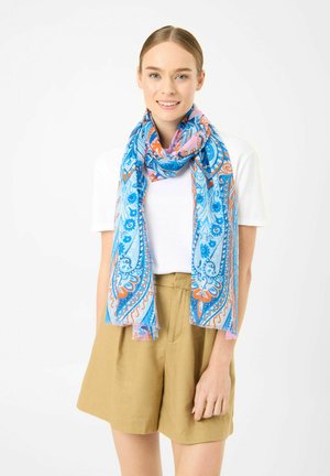 Young woman wearing a white t-shirt, tan high-waisted shorts, and a blue, orange, and pink patterned scarf draped around her neck.