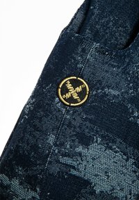 Tela de mezclilla en azul oscuro con un patrón texturizado. Presenta una insignia circular dorada cosida en el bolsillo.