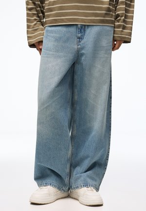Personne portant un jean bleu clair à jambes larges, des baskets blanches et une chemise à manches longues rayée marron et beige, debout avec les mains détendues.