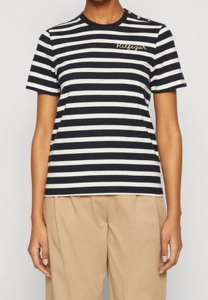 Vrouw die een zwart-wit horizontaal gestreept shirt met korte mouwen draagt met gouden knopen op de schouder en beige broek, staand met ontspannen armen.