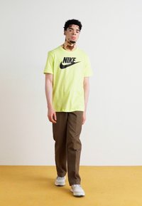 T-shirt Nike amarelo néon brilhante com logotipo preto, combinado com calças castanhas e ténis brancos, em pé sobre um chão amarelo contra uma parede neutra.