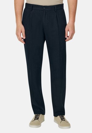 Schwarze, maßgeschneiderte Herrenhose mit vorderen Falten, getragen mit beigen Turnschuhen und einem beigen, eingesteckten Hemd vor einem einfarbigen Hintergrund.