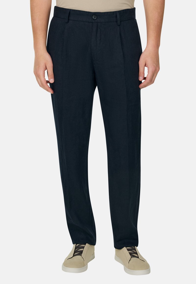 Pantalon noir pour homme, cintré avec plis sur le devant, porté avec des baskets beiges et une chemise beige rentrée, sur un fond uni.