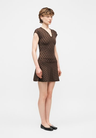 Νεαρή γυναίκα που στέκεται, φορώντας αμάνικο τοπ και φούστα με καφέ σχέδια και μαύρα επίπεδα παπούτσια, μπροστά σε απλό ανοιχτό φόντο.