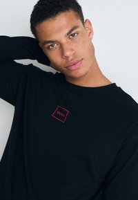 Cămașă neagră cu mânecă lungă din bumbac, având un mic logo rectangular roșu cu "HUGO" pe piept, design simplu și textură moale.