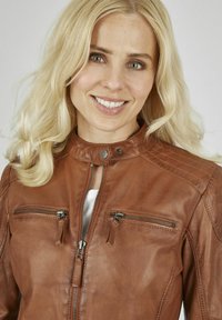 Braune Lederjacke mit einem Reißverschluss vorne, zwei mit Reißverschluss versehenen Brusttaschen und Stehkragen. Sie zeichnet sich durch eine glatte Textur und subtile Nähedetails aus.