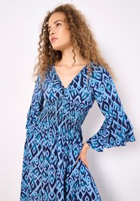 Blaue gemusterte Kleid mit V-Ausschnitt, geraffter Taille und ausgestellten Ärmeln. Das Design weist einen abstrakten Druck in verschiedenen Blautönen auf.