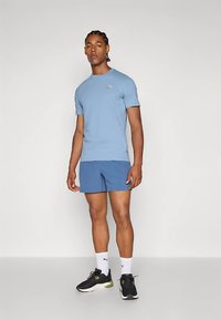 Camiseta de manga corta color azul claro con un logo sutil, combinada con pantalones cortos azul más oscuro. Zapatillas negras con detalles en lima y calcetines blancos.