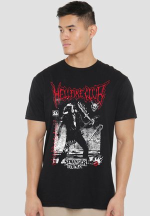 Schwarzes T-Shirt mit einer Grafik in Rot und Weiß, die eine Figur beim Gitarrespielen zeigt, mit den Schriftzügen "HELLFIRE CLUB" und "STRANGER THINGS".