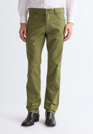 Mand iført olivengrønne bukser med lige ben, sorte læderstøvler og en hvid langærmet skjorte står mod en hvid baggrund.