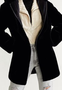 Zwarte, pluchen jas met ritssluiting en capuchon, met een gladde afwerking. Draagt over een crèmekleurige kabelgebreide trui en lichtgrijze distressed jeans.