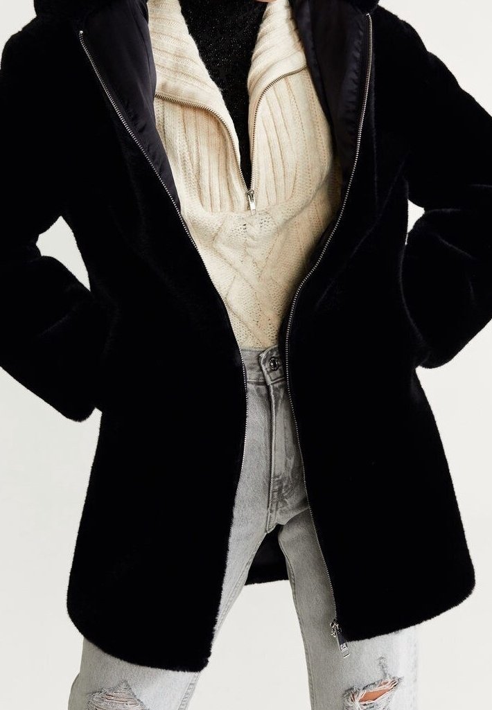 Zwarte, pluchen jas met ritssluiting en capuchon, met een gladde afwerking. Draagt over een crèmekleurige kabelgebreide trui en lichtgrijze distressed jeans.
