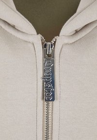 Light beige hoodie features a full-length silver zipper with a decorative pull tab and textured fabric for a soft finish.
