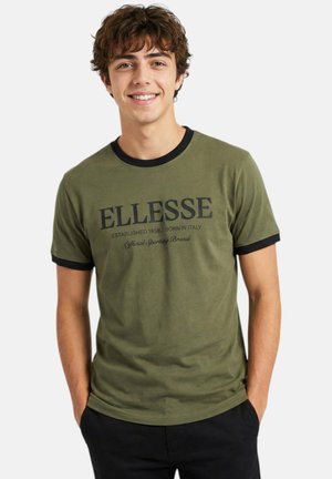 Jeune homme souriant portant un t-shirt Ellesse vert olive avec une bordure noire et les mains dans les poches sur un fond blanc.