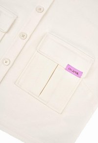 Beige knapp-up skjorta med två framfickor, en med en rosa etikett. Materialet verkar mjukt med en jämn textur.