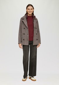Dubbelzijdige herringbone jas in grijs en bordeauxrood, met een grote kraag, zwarte knopen en een relaxte pasvorm, gedragen over een rode top en donkere broek.