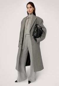 Szary wełniany płaszcz na szarym swetrze i szerokich spodniach z szerokimi nogawkami. Pod spodem koszula w paski. Czarna skórzana torba i czarne szpiczaste szpilki. Gładkie tekstury.