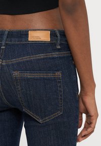 Vaqueros de mezclilla azul oscuro con un diseño clásico de cinco bolsillos, costuras naranjas y una etiqueta rectangular de cuero en la cintura.