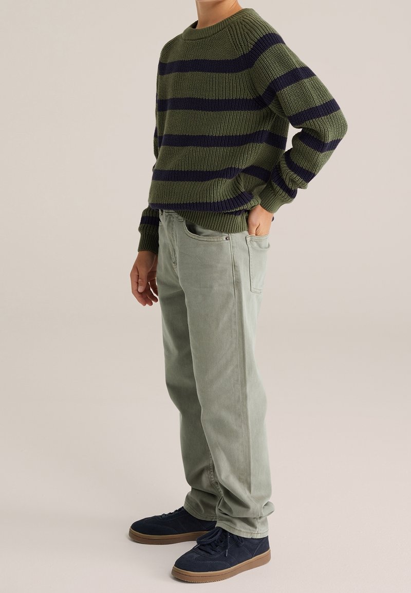 Personne portant un pull rayé olive et bleu marine, un pantalon vert clair et des baskets bleu foncé, debout avec une main dans la poche.