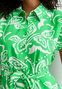 Vihreä napitettava mekko, jossa on valkoinen lehväkuvio, lyhyet hihat, kaulus ja sidottu vyötärö. Laadukas kangas, jossa on hieman kiiltävä pinta.