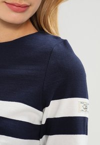 Pull rayé bleu marine et blanc avec un col rond, en coton doux, et une étiquette sur la manche gauche affichant le logo de la marque.