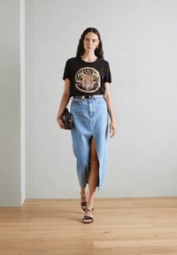 T-shirt nera con design grafico circolare, abbinata a una gonna di jeans azzurri alta in vita con spacco e sandali neri con cinturino.