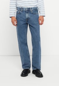 Persona que lleva puestos unos jeans rectos azules, mocasines negros y una camisa blanca con rayas horizontales en azul claro, de pie contra un fondo blanco liso.