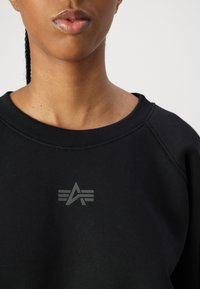 Svart hettegenser med rund hals, med en subtil grå logo på brystet. Stoffet har en glatt tekstur. Ermehullene er i raglanstil.